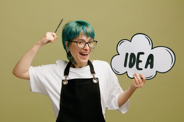 Excitée jeune femme barbier portant des lunettes uniforme montrant une bulle d'idée regardant la caméra tenant des ciseaux près de la tête isolé sur fond vert olive