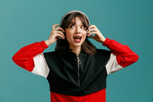 Excitée jeune femme caucasienne portant des écouteurs regardant sur le côté tout en saisissant des écouteurs et en écoutant de la musique isolée sur fond bleu