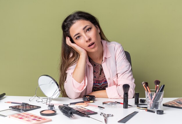 Excitée jeune fille brune assise à table avec des outils de maquillage