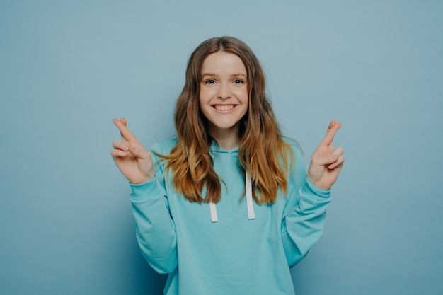 Excitée jeune fille souriante aux cheveux ondulés ombrés en gardant les doigts croisés en regardant la caméra portant un pull décontracté posant isolé sur fond de studio bleu clair. concept de langage corporel