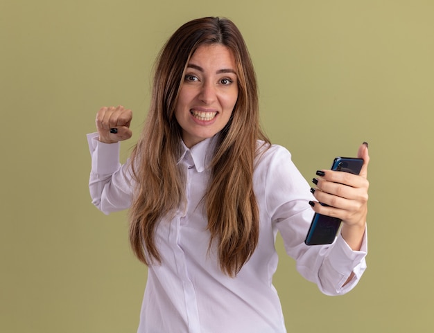 Excitée jeune jolie fille caucasienne garde le poing et tient le téléphone isolé sur un mur vert olive avec espace de copie