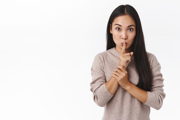 Excitée mignonne petite amie est-asiatique prépare la surprise de la Saint-Valentin en disant à un ami des idées demandant de ne pas dire de se taire, appuyez sur le doigt sur les lèvres pliées dans un geste de silence et regardez la caméra