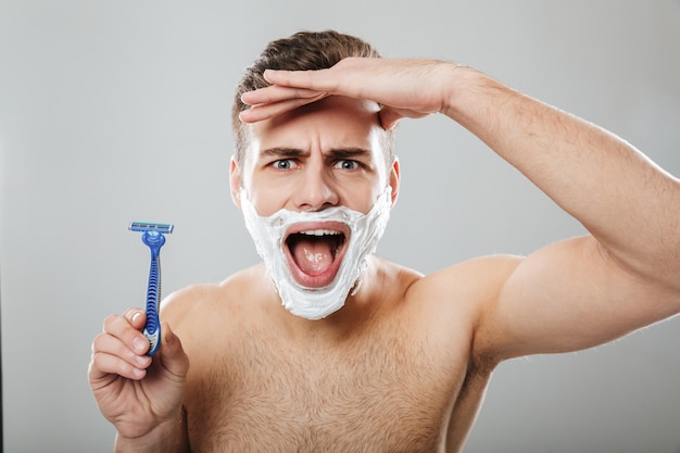 Photo gratuite expressions faciales drôles d'un homme non rasé faisant la procédure du matin avec un rasoir et de la crème sur le visage sur un mur gris