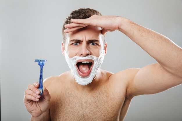 Expressions faciales drôles d'un homme non rasé faisant la procédure du matin avec un rasoir et de la crème sur le visage sur un mur gris