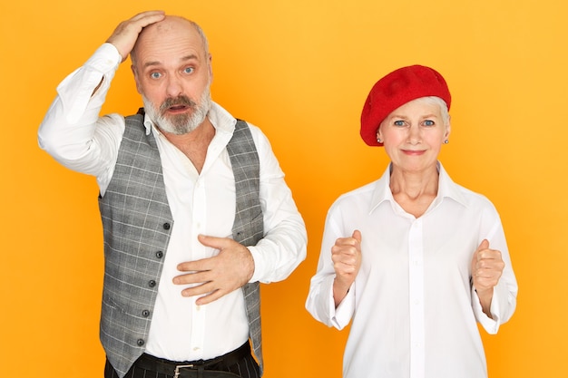 Expressions faciales humaines et langage corporel. Image isolée de retraité homme barbu frustré touchant sa tête chauve ayant regard perplexe, femme mûre d'espoir en bonnet rouge serrant les poings