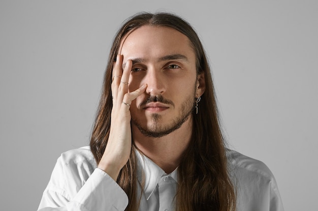 Expressions faciales humaines. Tir isolé de beau jeune homme de race blanche mal rasé aux cheveux longs posant, ayant un regard sérieux, tenant la main sur son visage, portant une chemise élégante et des accessoires