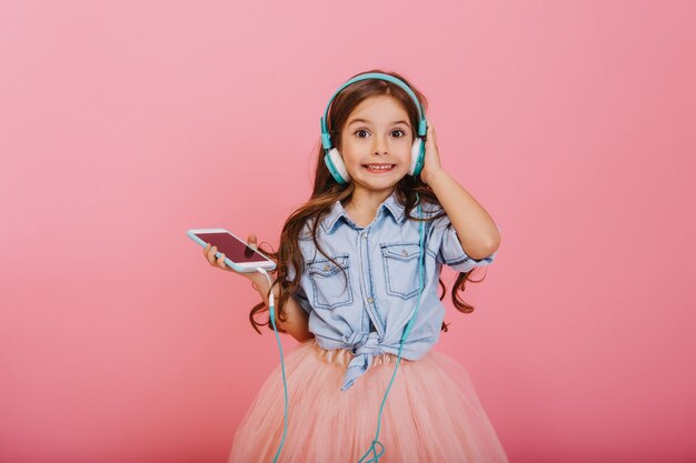 Exprimant la positivité d'un enfant heureux, écoutant de la musique avec des écouteurs bleus isolés sur fond rose. Jolie petite fille aux longs cheveux brune souriant à la caméra en jupe en tulle