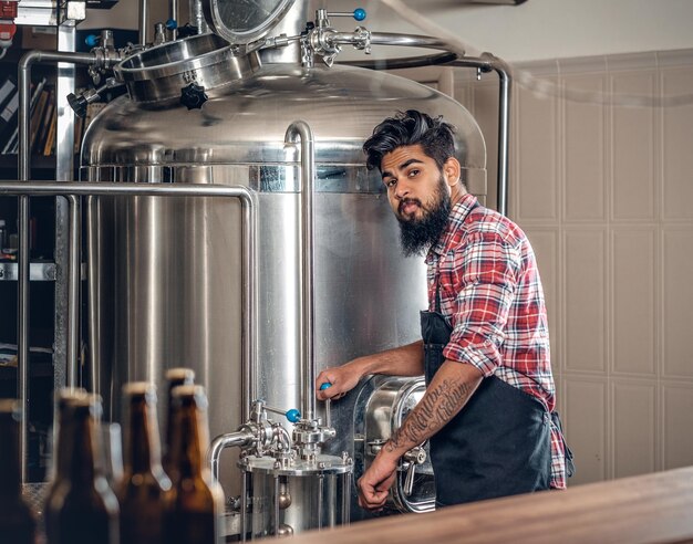 Fabricant masculin hipster barbu tatoué indien présentant de la bière artisanale dans la microbrasserie.