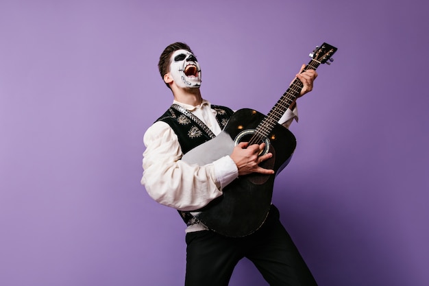 Face art guy s'imagine musicien de rock et pose avec émotion à la guitare. Portrait intérieur de l'homme sur mur lilas.