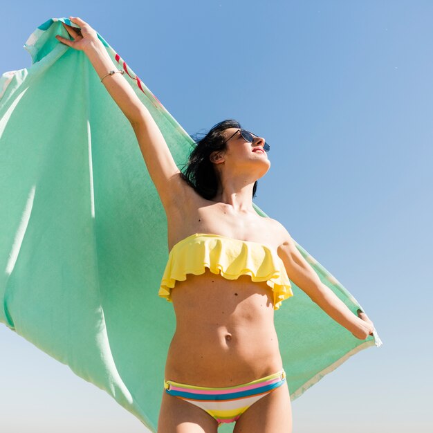 Faible angle de vue de la jeune femme en bikini tenant une serviette à la main, debout contre le ciel bleu