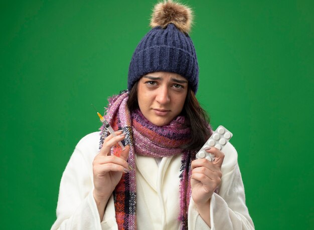 Faible jeune femme malade portant chapeau d'hiver robe et écharpe tenant la seringue et pack de comprimés à l'avant isolé sur mur vert
