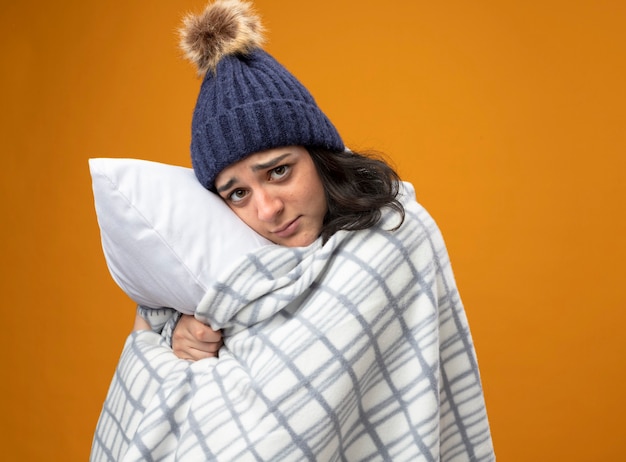 Faible jeune femme malade portant robe chapeau d'hiver enveloppé de plaid debout en vue de profil étreindre oreiller à l'avant isolé sur mur orange