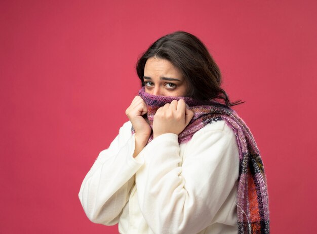 Faible jeune femme malade portant robe et écharpe debout en vue de profil couvrant la bouche avec un foulard à l'avant en gardant les mains sur l'écharpe isolé sur mur rose