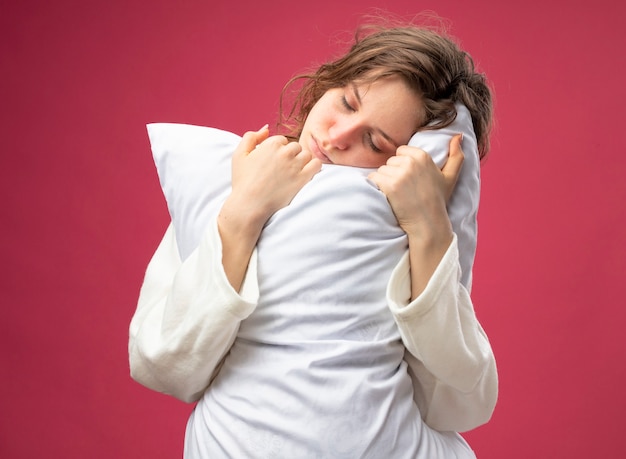 Faible jeune fille malade aux yeux fermés portant une robe blanche oreiller étreint isolé sur rose