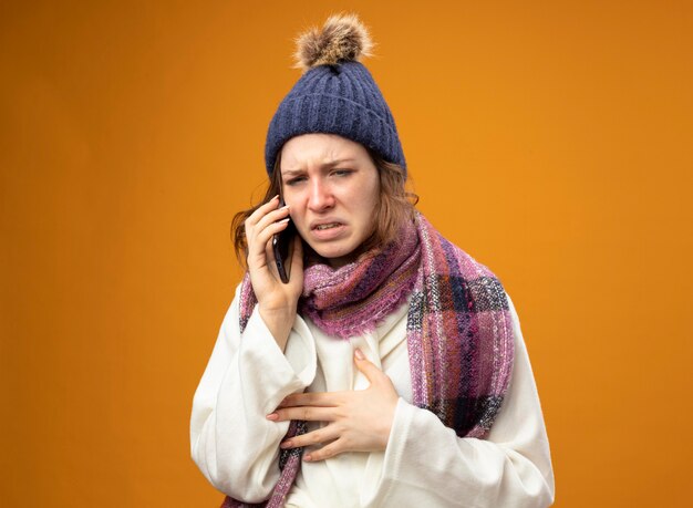 Faible jeune fille malade portant robe blanche et chapeau d'hiver avec écharpe parle au téléphone isolé sur orange