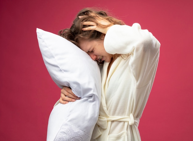 Faible jeune fille malade portant robe blanche oreiller étreint mettant la main sur la tête douloureuse isolé sur rose