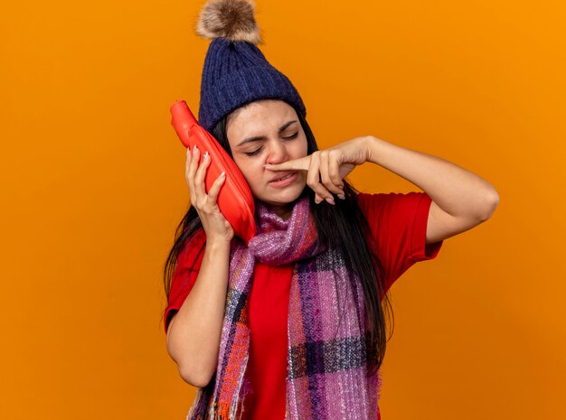 Faible jeune fille malade de race blanche portant un chapeau d'hiver et une écharpe touchant le visage avec un sac d'eau chaude essuyant le nez avec le doigt avec les yeux fermés isolé sur un mur orange avec espace de copie