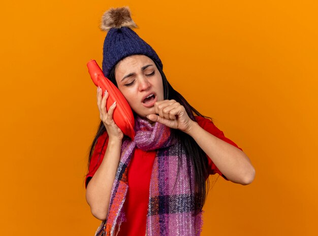 Faible jeune fille malade de race blanche portant un chapeau d'hiver et une écharpe touchant le visage avec un sac d'eau chaude toux en gardant le poing près de la bouche avec les yeux fermés isolé sur un mur orange avec espace de copie