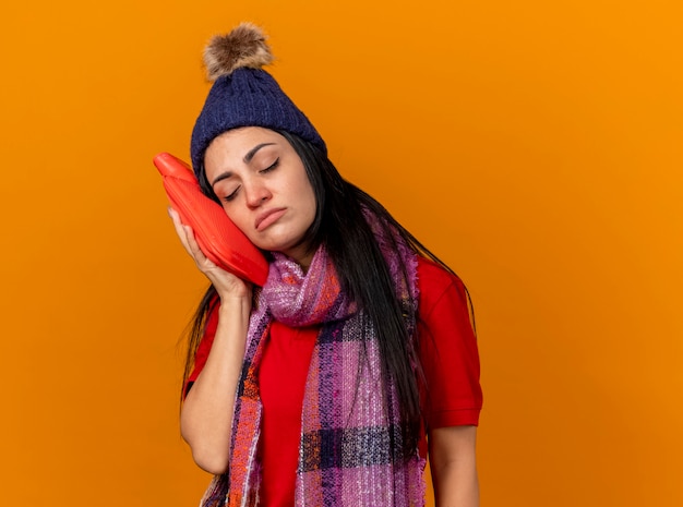 Faible jeune fille malade de race blanche portant un chapeau d'hiver et une écharpe touchant le visage avec un sac d'eau chaude avec les yeux fermés isolé sur fond orange avec copie espace