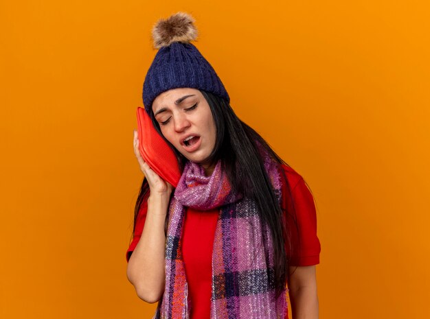 Faible jeune fille malade de race blanche portant un chapeau d'hiver et une écharpe touchant le visage avec un sac d'eau chaude avec les yeux fermés isolé sur un mur orange avec copie espace