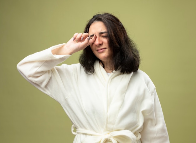 Faible jeune fille malade de race blanche portant robe essuyant les yeux avec les yeux fermés isolé sur fond vert olive