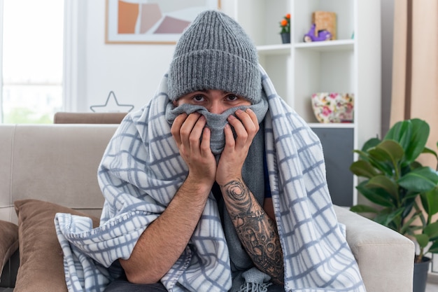 Faible jeune homme malade portant une écharpe et un chapeau d'hiver assis sur un canapé dans le salon enveloppé dans une couverture regardant la caméra couvrant la bouche avec une écharpe