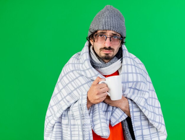 Faible jeune homme malade portant des lunettes chapeau d'hiver et écharpe enveloppé dans un plaid tenant une tasse de thé saisissant plaid à l'avant isolé sur mur vert