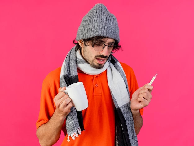 Faible Jeune Homme Malade Portant Des Lunettes Chapeau D'hiver Et écharpe Tenant Tasse Et Thermomètre Regardant Vers Le Bas Isolé Sur Mur Rose