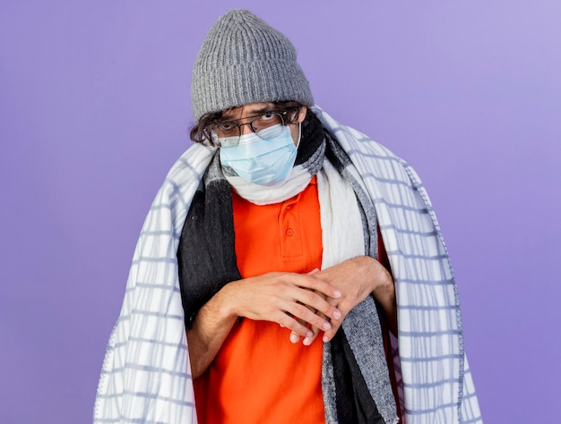 Photo gratuite faible jeune homme malade de race blanche portant des lunettes avec chapeau d'hiver masque et écharpe enveloppé de plaid en gardant les mains ensemble isolé sur mur violet