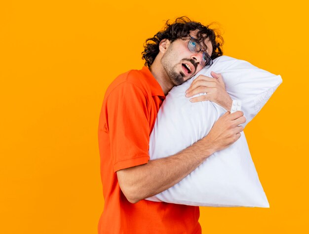 Photo gratuite faible jeune homme malade de race blanche portant des lunettes tenant oreiller et pack de comprimés médicaux mettant la tête sur l'oreiller à tout droit isolé sur fond orange avec copie espace