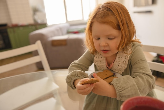 Fairskinned petite fille enfant aux cheveux rouges en robe snack alors qu'il était assis à table pendant la journée Concept de nutrition