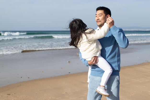 Famille asiatique amusée marchant ensemble sur la plage. Père et fille en vêtements décontractés dansant près de l'eau et riant. Convivialité, amour, concept parental