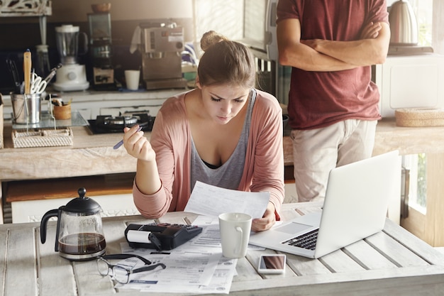 Famille caucasienne ayant du mal à payer les factures. Jeune femme lisant un document avec un regard concentré, tenant un stylo, calcul des finances à la maison