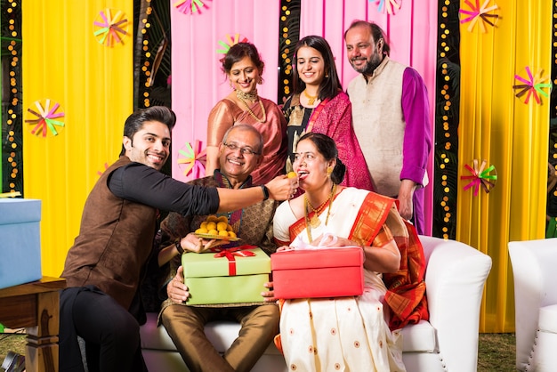 Famille indienne multigénérationnelle mangeant des bonbons tout en célébrant un festival ou une occasion vêtue de vêtements traditionnels, assise sur un canapé ou un canapé