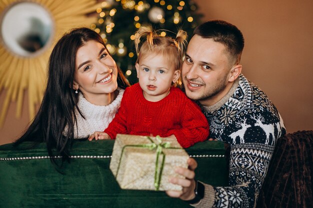 Famille avec petite fille assise devant un arbre de Noël et une boîte-cadeau déballée