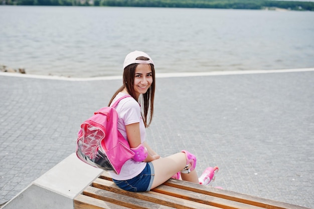 Fantastique jeune femme en tenue décontractée et casquette assise sur le banc du skatepark avec des patins à roues alignées