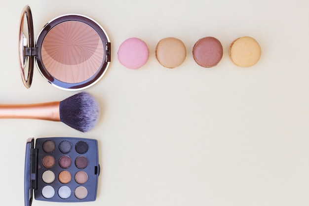 Photo gratuite fard à joues avec macaron; pinceau de maquillage et palette de fard à paupières sur fond beige