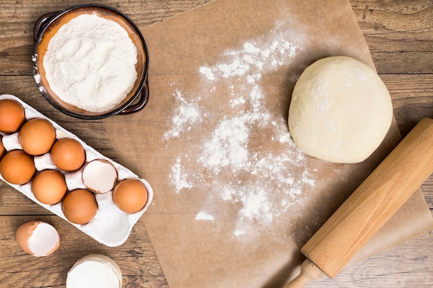 Farine; carton d&#39;oeufs; pâte; rouleau à pâtisserie sur papier parchemin sur le bureau en bois