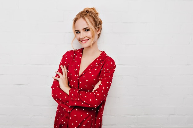 Fascinante fille bouclée porte un pyjama rouge posant avec un sourire timide. portrait de femme effrayante debout avec les bras croisés sur le mur blanc.