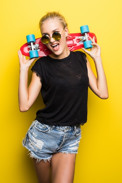 Fashion happy smiling hipster cool girl dans des lunettes de soleil et des vêtements colorés avec skateboard s'amuser à l'extérieur sur le fond orange