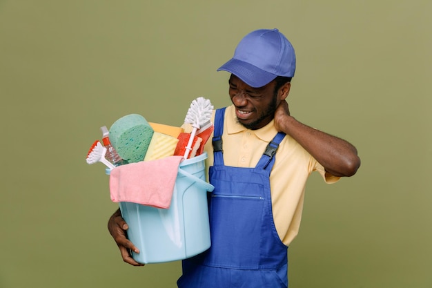 Photo gratuite fatigué attrapé le cou tenant un seau d'outils de nettoyage jeune homme nettoyant afro-américain en uniforme avec des gants isolés sur fond vert