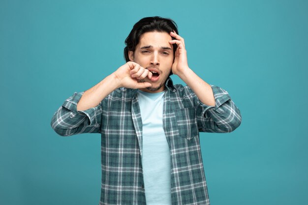 fatigué jeune bel homme gardant les mains sur la tête et près de la bouche regardant la caméra bâillant isolé sur fond bleu