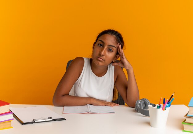 Fatigué de jeune écolière assis au bureau avec des outils scolaires mettant la main sur la tête sur orange