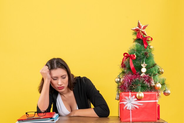 Fatigué de jeune femme en costume près de sapin de Noël décoré au bureau sur jaune