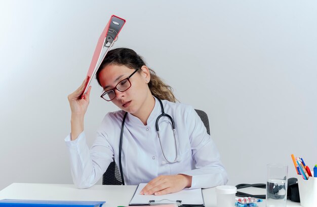 Fatigué de jeune femme médecin portant une robe médicale et un stéthoscope et des lunettes assis au bureau avec des outils médicaux touchant la tête avec le dossier avec les yeux fermés isolés