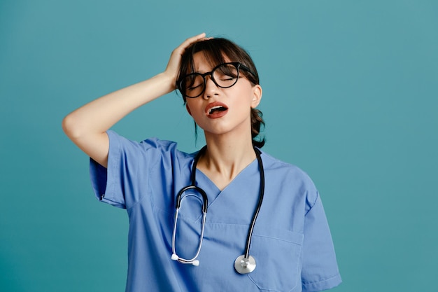 Fatigué jeune femme médecin portant un stéthoscope fith uniforme isolé sur fond bleu
