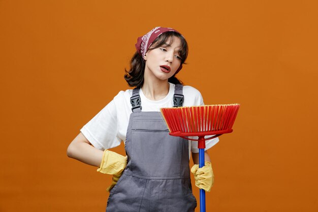 Fatigué jeune femme nettoyeuse portant des gants en caoutchouc uniformes et un bandana tenant une vadrouille à raclette en le regardant tout en gardant la main sur la taille isolée sur fond orange
