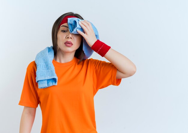 Fatigué de jeune femme sportive portant un bandeau et des bracelets avec une serviette autour du cou regardant vers le bas essuyant la sueur avec une serviette