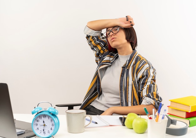 Fatigué de jeune fille étudiante portant des lunettes assis au bureau avec des outils universitaires mettant le bras sur le front avec les yeux fermés isolé sur fond blanc