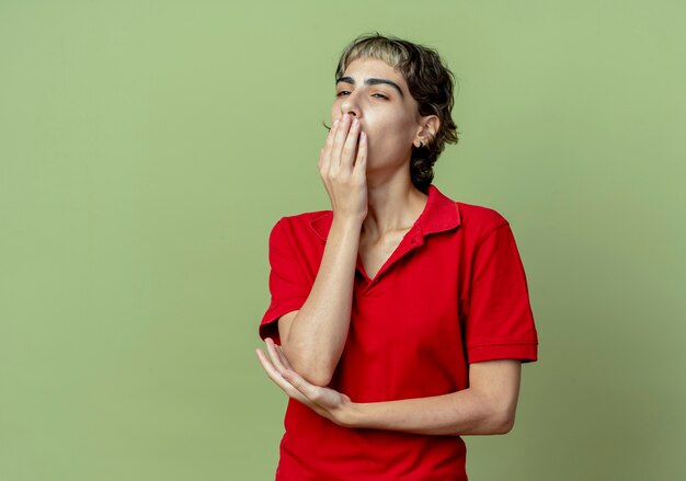 Fatigué de jeune fille de race blanche avec coupe de cheveux de lutin regardant le côté bâillant avec les mains sur la bouche et sous le coude isolé sur fond vert olive avec espace de copie